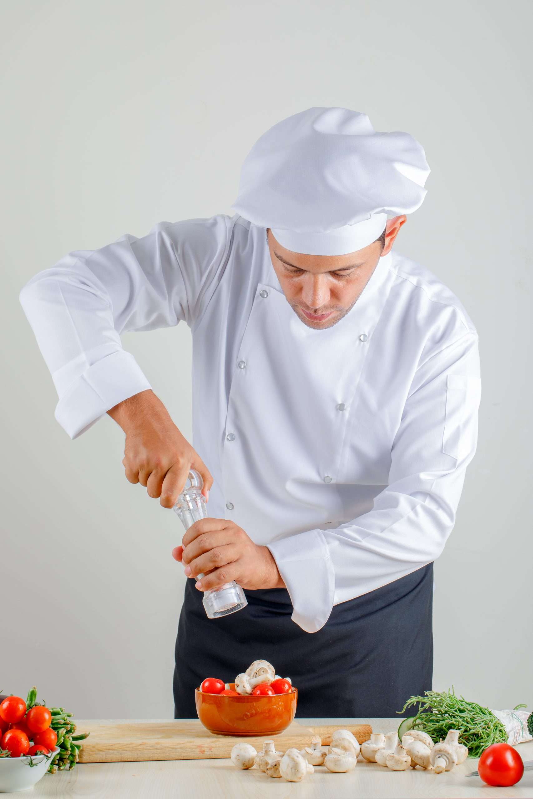 Male chef adding salt into food in uniform, hat and apron in kitchen , front view.